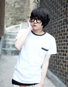 a young man wearing glasses talking on a cell phone while standing next to a brick wall