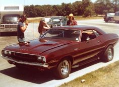 two people standing next to a red muscle car