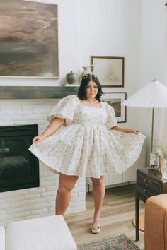 a woman standing in front of a fire place wearing a white dress with floral print