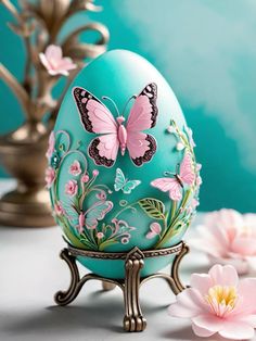 an egg with butterflies painted on it sitting next to pink flowers and a gold vase