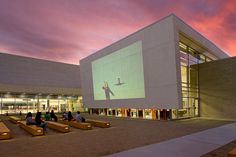 people are sitting on benches in front of a building with a large screen projected on it