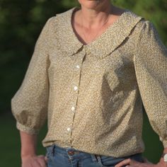 a woman wearing a blouse and jeans standing in front of some trees with her hands on her hips