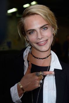 a woman with blonde hair and piercings smiles at the camera while pointing to her right