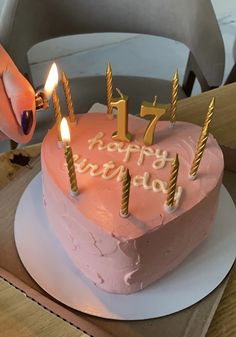 a pink birthday cake with candles on it and someone lighting the candle in the shape of a heart