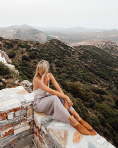 a woman sitting on top of a stone wall next to a lush green hillside covered in trees