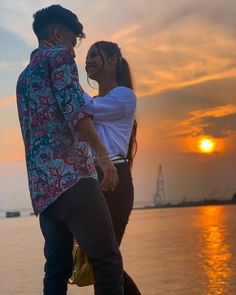 a man and woman standing next to each other near the water at sunset or sunrise