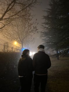 two people walking down a sidewalk at night in the fog with street lights behind them