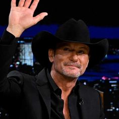 a man in a black suit and cowboy hat waves to the crowd while sitting down