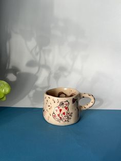 a ceramic cup with an image of a cat painted on it sits in front of a green plant