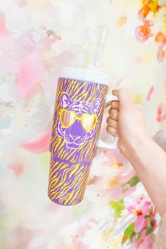 a person holding a cup with a purple tiger design on it and yellow sunglasses in their hand