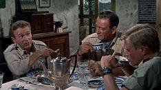 three men sitting at a table with plates and cups in front of them, eating