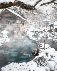 two people swimming in a hot spring surrounded by snow covered rocks and trees, with a cabin on the other side