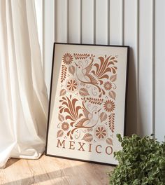 a framed mexican art piece sitting on top of a wooden floor next to a potted plant