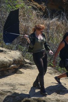 a woman holding an umbrella while walking down a dirt road next to a man with a backpack