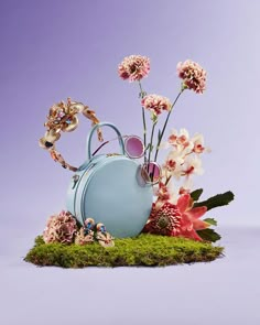 a teapot with flowers and sunglasses on top of mossy ground in front of a purple background