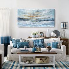 a living room filled with white furniture and blue pillows on top of a table in front of a painting