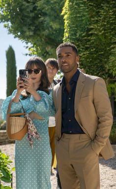 a man and woman standing next to each other in front of some trees, taking pictures with their cell phones