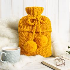 a yellow knitted bag with pom - poms next to a cup of coffee