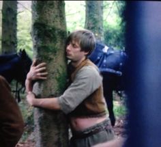 a man hugging a tree in the woods with two horses behind him and another person standing next to it