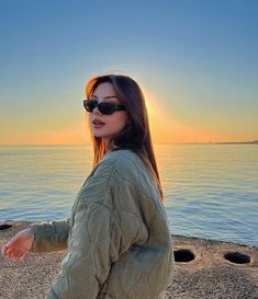 a woman in sunglasses standing on the beach at sunset with her back to the camera