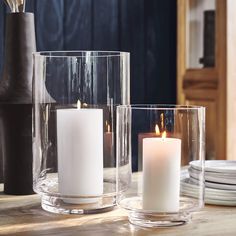 two clear vases filled with white candles on top of a wooden table next to plates