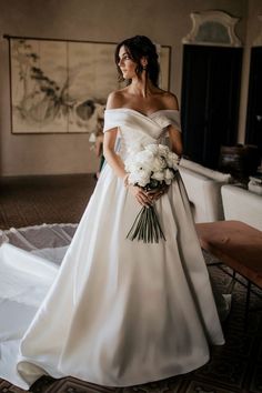 a woman in a white wedding dress standing on a bed holding a bouquet of flowers