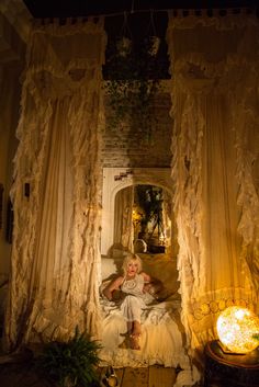 a woman is sitting on a bed in a room with curtains and lights around her