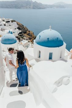 a man and woman standing on top of a white building next to the ocean with blue domes