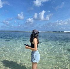 a woman standing in shallow water holding a cell phone and looking at the camera with an island in the background