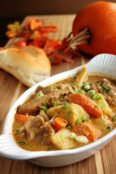 a white bowl filled with stew next to bread on a wooden table and an orange pumpkin in the background