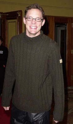 a man wearing glasses and a brown sweater smiles at the camera while standing in a hallway