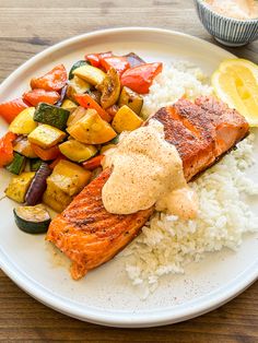 a white plate topped with salmon, rice and veggies next to a cup of sauce