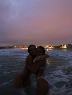 two women hugging each other in the water at night on a beach with buildings in the background