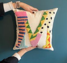 two hands holding a decorative pillow that has the letter v on it and is decorated with multicolored yarn