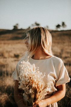 a woman standing in the middle of a field holding a bunch of dried flowers on her shoulder