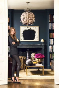 a woman standing in front of a fireplace with a chandelier hanging from the ceiling