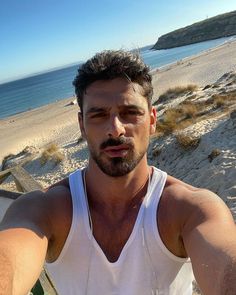 a man in white tank top standing on beach next to water and sand with his arms crossed