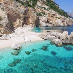 the beach is surrounded by large rocks and clear blue water, as well as cliffs