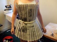 a woman in a dress made out of newspaper strips is standing next to a table