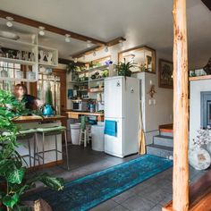 a woman is standing in the kitchen next to a refrigerator and table with potted plants on it