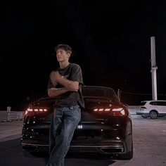 a man leaning on the hood of a black car in a parking lot at night