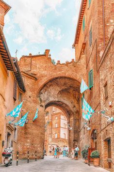 people are walking through an alleyway with flags flying in the air and brick buildings on either side