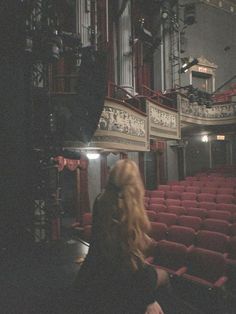 a woman sitting in an empty theater looking at the stage with red seats on either side