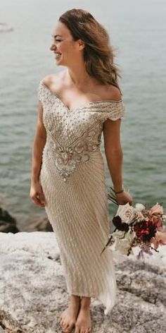 a woman in a dress is walking on the rocks by the water with her bouquet