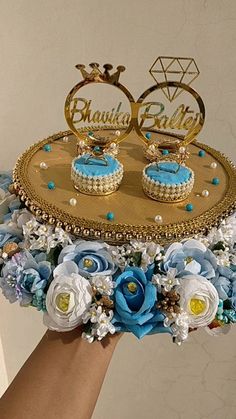 a hand holding a cake decorated with blue flowers and gold rings on top of it