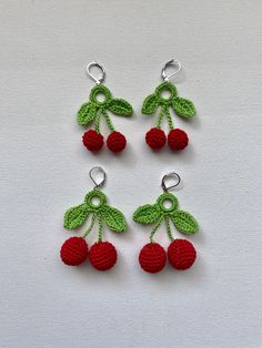 four crocheted cherries hanging from hooks on a white surface with green leaves and red berries