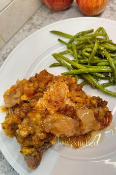 a white plate topped with green beans and meat covered in sauce next to an apple