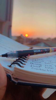 a person holding a notebook with a pen on top of it and the sun setting in the background