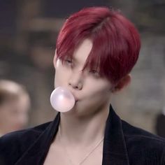 a man with red hair blowing bubbles in his mouth while wearing a black jacket and silver necklace