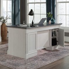 an office desk with a laptop on it in front of two windows and a rug